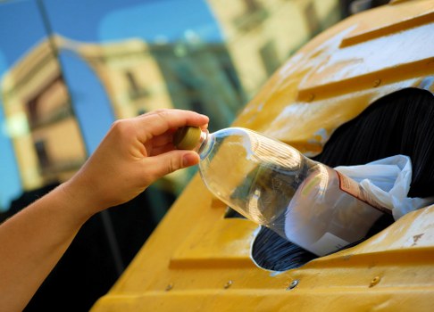 A person's hand, wearing a beige glove, is placing a clear plastic bottle into a black circular opening on the yellow exterior of a waste collection vehicle, likely for rubbish or recycling collection. The bottle appears to be a transparent plastic container with a brown cap, containing some residual liquid. The vehicle's body is made of shiny, painted metal with a smooth finish, showing reflections of nearby buildings and the sky, which suggests it is outdoors on a sunny day. In the background, blurred structures or buildings can be seen, indicating an urban or suburban environment. This scene is typical of rubbish collection or waste disposal operations, with the vehicle positioned on a street or driveway. The image relates to waste management services offered by companies such as Office Clearance Brimsdown, providing professional rubbish removal in the local area close to Brimsdown, Essex, or nearby postcode regions.
