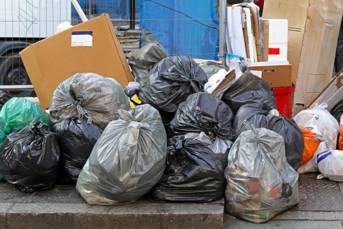 A collection of black and grey plastic rubbish bags filled with waste, piled on a paved surface outside a building. Some bags are more tightly packed and knotted at the top, while others are loosely gathered, showing hints of their contents. The bags are arranged irregularly, with a few partially overlapping each other. In the background, there are cardboard boxes, wooden panels, and various other discarded items, indicating an area of waste collection or clearance outside a commercial or residential property. The environment appears urban with a concrete curb and a slight elevation difference from the street or pathway. This scene illustrates typical rubbish disposal and clearance activity that a company like Office Clearance Brimsdown would handle in the local postcode area, supporting professional rubbish removal services in the vicinity of Brimsdown.