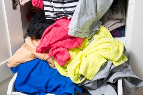 A close-up view of an open white wardrobe drawer filled with a disorganized pile of various clothing items, including a beige pair of trousers, a bright pink top, a yellow shirt, a blue garment, and several other pieces in different colors and fabrics. The clothes appear to be casual wear made from materials like cotton and polyester, with some folded and others crumpled, occupying most of the visible space in the drawer. Surrounding the drawer are other sections of the wardrobe with additional clothes and storage compartments, set against a neutral background. This scene reflects typical household laundry or decluttering activities related to rubbish removal or clearance services such as those offered by Office Clearance Brimsdown, which assist in clearing out accumulated personal or office clothing and textile waste in the local area near Brimsdown, Essex.