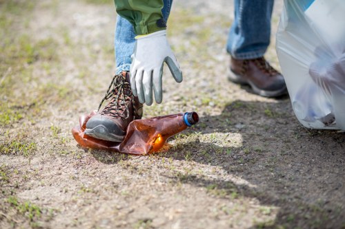 A person wearing white work gloves and blue jeans is seen picking up a brown glass bottle from a patch of grass and dirt, with an outdoor environment visible in the background. The individual is standing next to a large, clear plastic garbage bag filled with rubbish, which is positioned on the ground beside them. The person's footwear includes sturdy brown work boots, and the bottle appears to be discarded litter. The scene suggests a cleanup activity, possibly part of a rubbish removal or environmental clearance service in Brimsdown, with the focus on removing trash from outdoor spaces to improve local surroundings. The natural outdoor lighting highlights the textures of the soil, grass, and the reflective surface of the glass bottle, emphasizing a scene of community or environmental effort to clear waste from a residential or semi-rural area around Brimsdown, with no other objects or structures visible. This detailed visual description supports the service category of rubbish removal offered by Office Clearance Brimsdown, based near the postcode region.