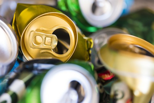 A close-up view of several crushed aluminum cans, predominantly in gold, silver, green, and blue colors, with textured metallic surfaces showing creases, dents, and pull-tab openings. The cans are piled together, some overlapping, in a heap likely intended for recycling or disposal, with a blurred background suggesting an indoor or outdoor setting. The focus is on the tops and bodies of the cans, highlighting their lightweight, crinkled frames and shiny finishes. This image relates to rubbish removal services offered by Office Clearance Brimsdown, emphasizing the collection and disposal of metal waste in a professional and environmentally conscious manner, suitable for a location within the postal code area serving Brimsdown. The detailed description ensures clear understanding for accessibility and supports local SEO relevance for waste management solutions in the area. The scene visually captures the types of debris handled during rubbish clearance in a typical urban or suburban environment, aligning with the services provided by the company.
