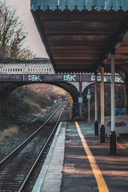 The image depicts an outdoor railway platform at dusk, with the perspective extending down the tracks towards a distant signal light. In the foreground, a protective canopy with a dark wooden roof and