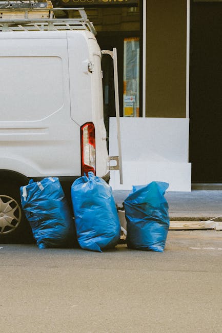 A small burgundy flatbed truck parked on an urban street in front of a modern glass-fronted office building. The truck's cargo area is filled with various types of rubbish, including large black and w