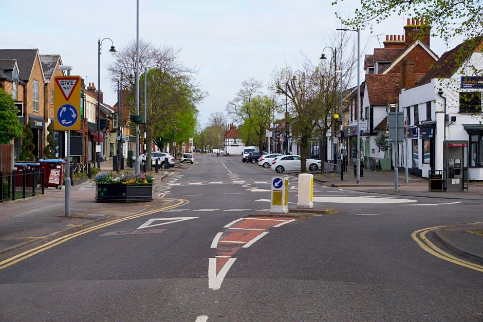A view of a small urban street intersection in a residential and commercial area during daylight hours. The scene includes a paved road with various road markings, including a central red and white st